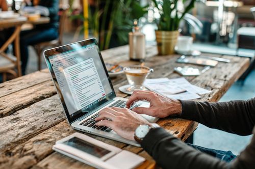 Hlavním objektem na obrázku je laptop umístěný na dřevěném stole, se kterým pracuje osoba v civilním oblečení.