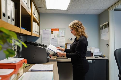 Žena stojí u kopírky v kanceláři a drží dokumenty. Kolem jsou police s pořadači a na stěnách visí papíry.