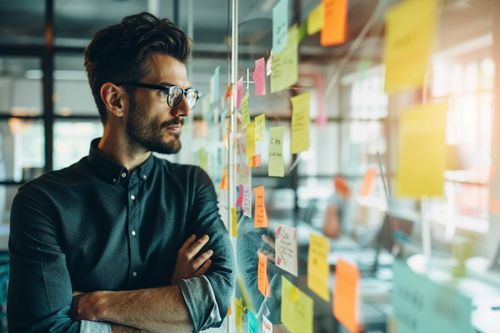 Muž s brýlemi a vousy stojí před skleněnou stěnou pokrytou barevnými poznámkovými bločky.