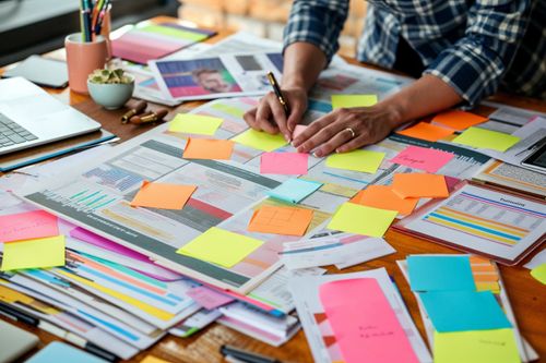 Stůl pokrytý barevnými poznámkovými bločky a tiskovými materiály s rukama osoby, která píše.