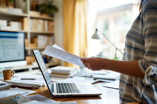 Osoba drží papír a pracuje na laptopu v domácím kancelářském prostředí.