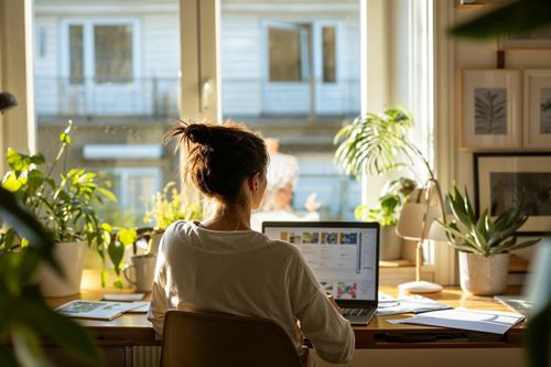 Žena sedí u stolu a pracuje na laptopu ve světlé místnosti s rostlinami.
